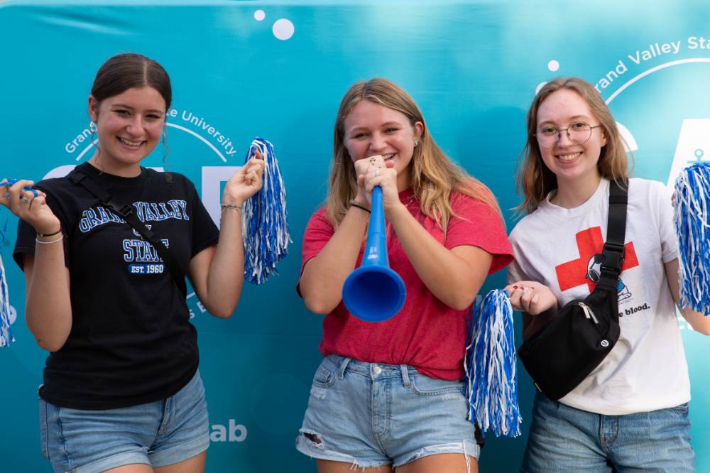 three students posing in front of CAB backdrop at Laker Kickoff photo booth smiling and blowing horn